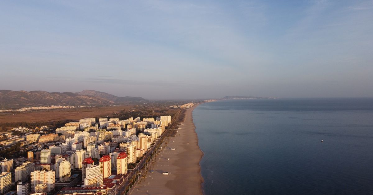 Vista aérea de una población costera mediterránea con playa y edificios al atardecer.